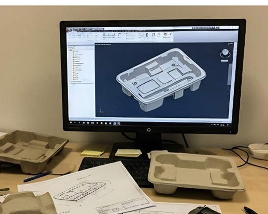 A cluttered engineering desk with a CAD monitor displaying a 3D mold design next to physical pulp packaging samples.